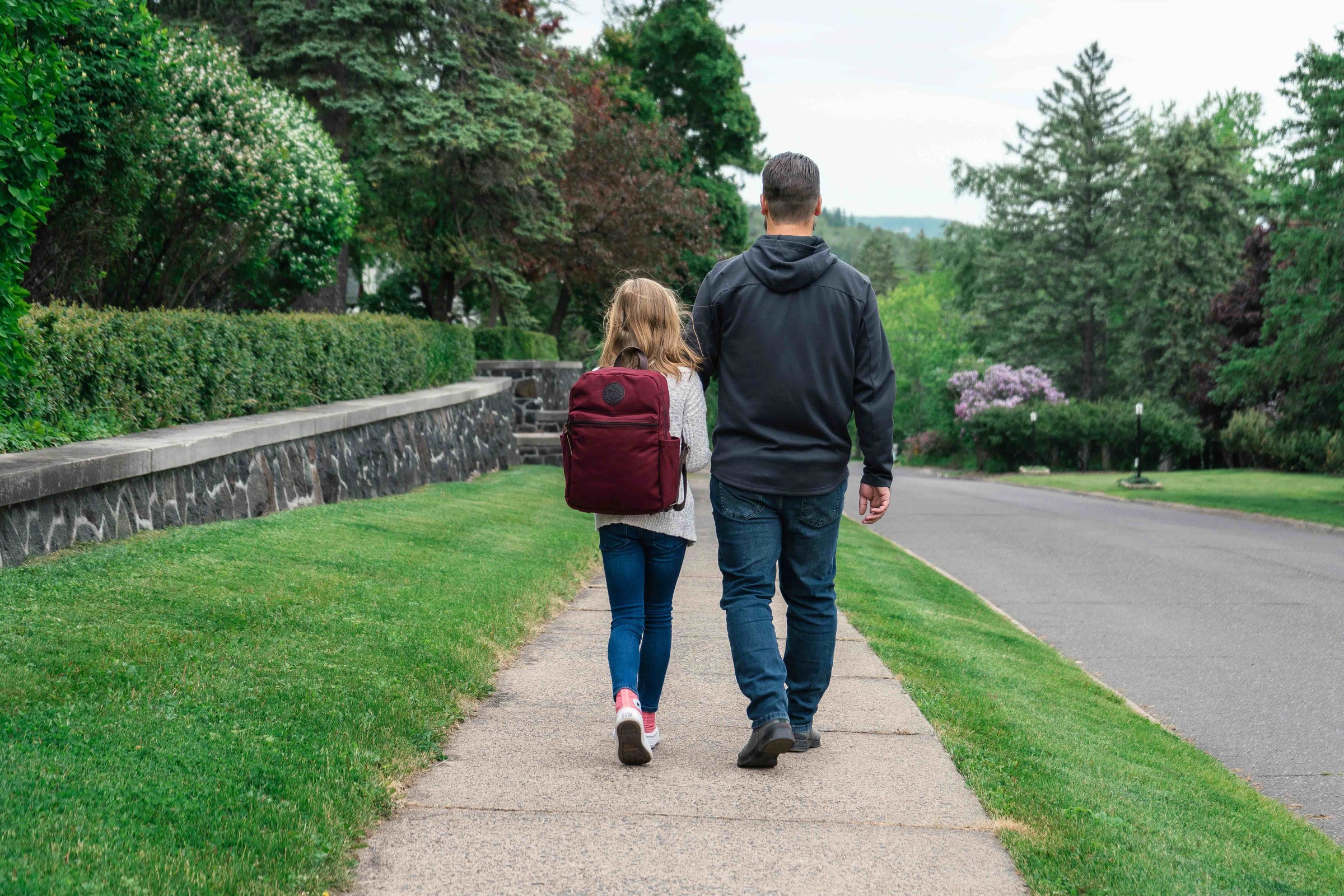 A dad and daughter walking down the street. The daughter is wearing a burgundy Standard Backpack by Duluth Pack for back to school.  
