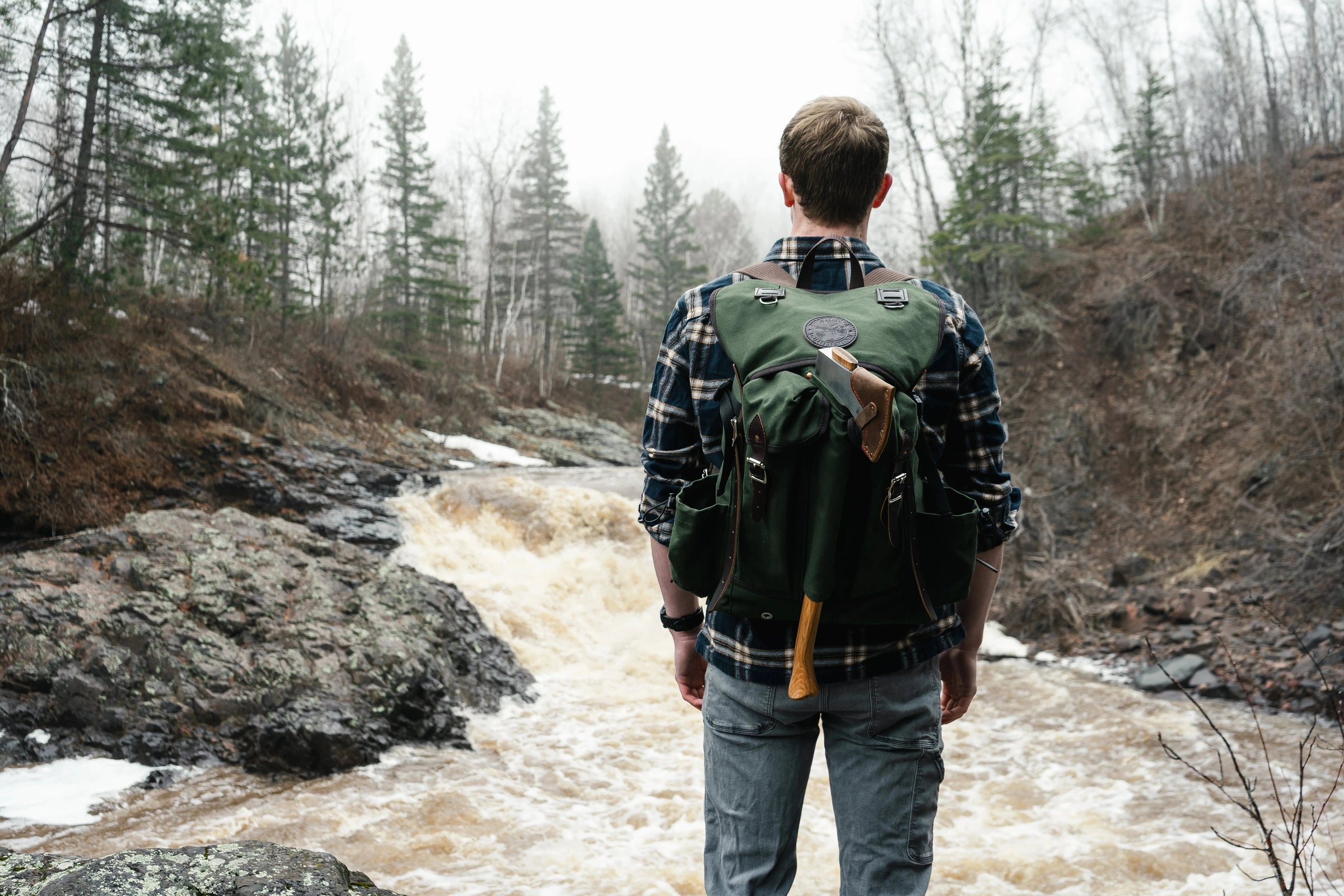 A man wearing an olive drab Duluth Pack Bushcrafter pack. The man is standing in front of a waterfall. 