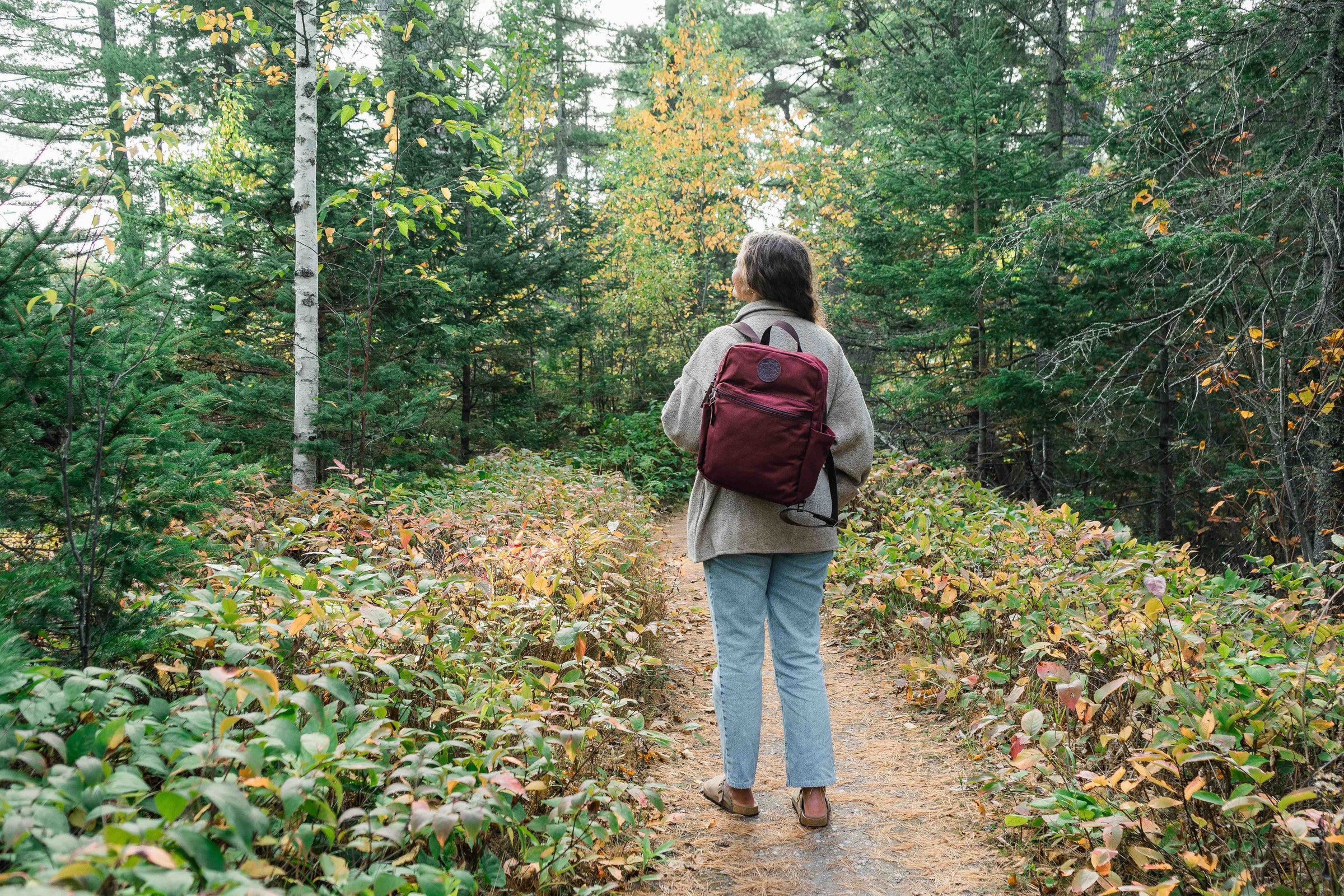 Woman in the woods wearing a burgundy Duluth Pack Standard Backpack with fall colored leaves in the background. 