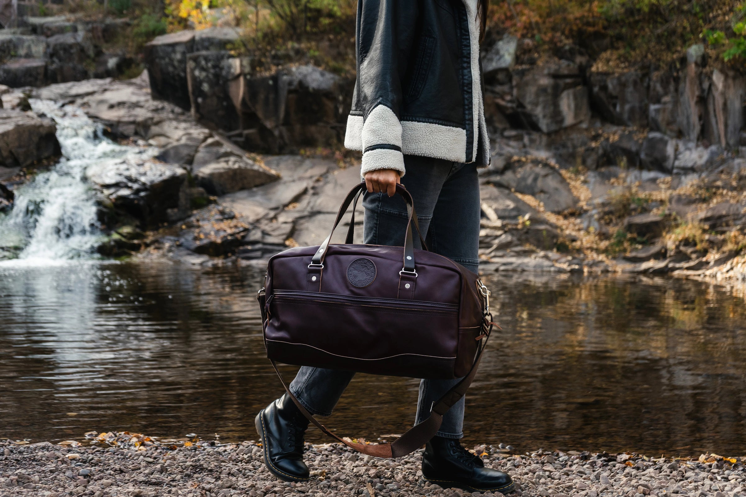 A woman walking on a rocky beach in a leather coat with a brown leather Duluth Pack Weekender Duffel. There is a waterfall in the background. 