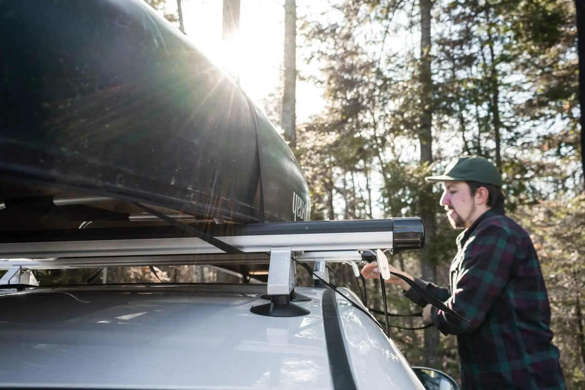A man wearing a Duluth Pack Flannel attaching a canoe to a Spring Creek Manufacturing Spring Creek DIY Suction Cup Rack Kit onto the man's car with a forest in the background. 