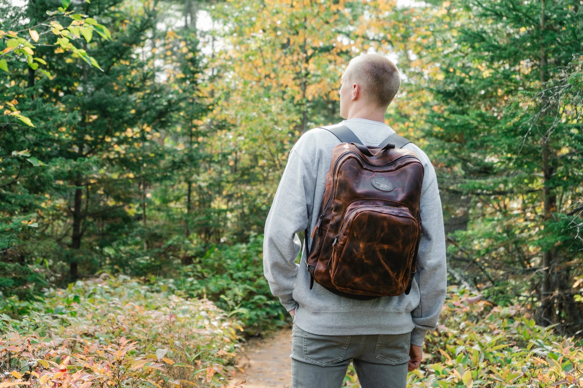 A man standing in a green forest wearing Duluth Pack Heritage Leather goods. 