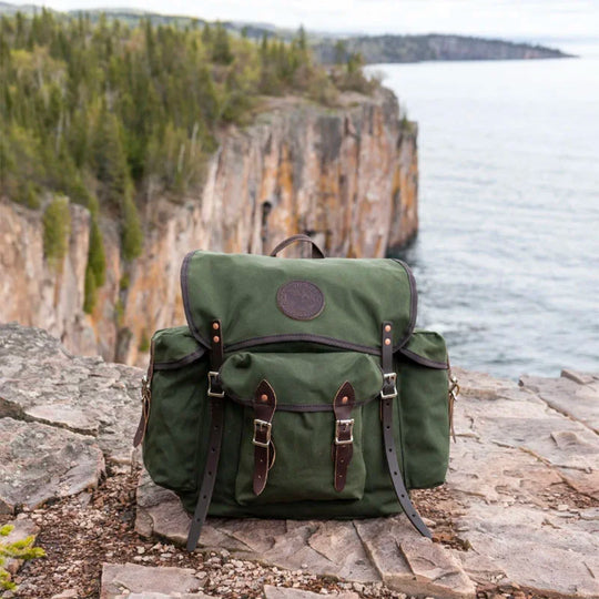 Wanderer backpack in Olive Drab canvas with leather reinforcements, showcased on a rugged cliffside overlooking a forested shoreline and lake.