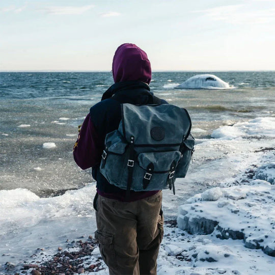 Wanderer backpack in waxed grey canvas with leather reinforcements, worn by a hiker overlooking a frozen lakeshore, showcasing durable outdoor gear.