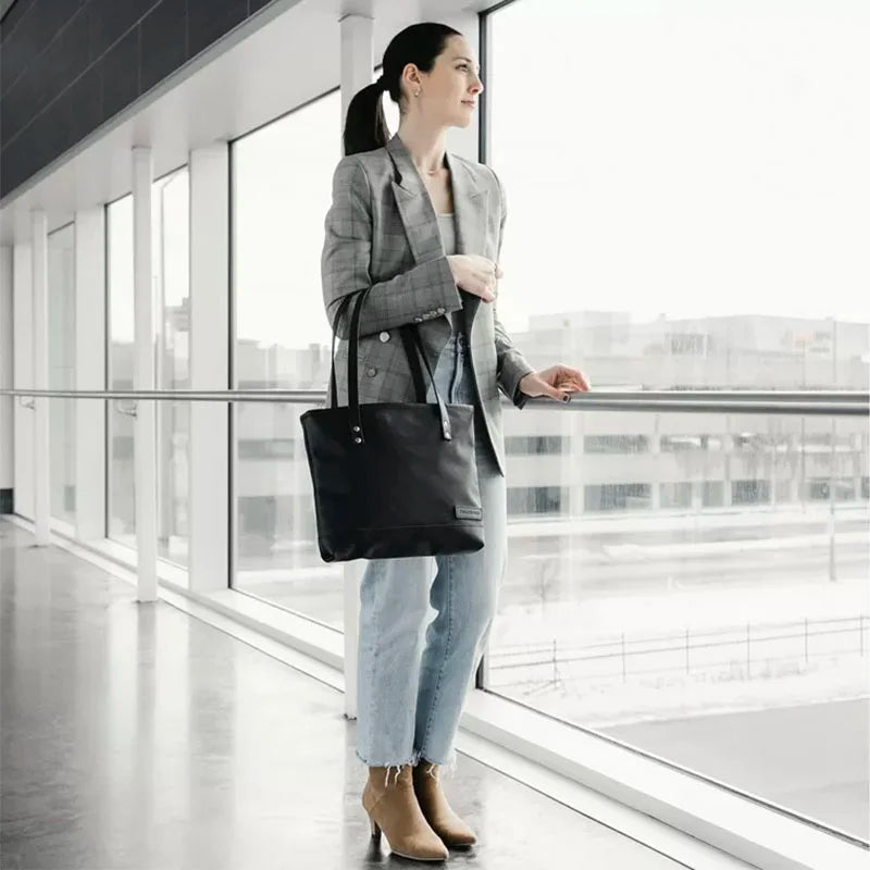 Superior Series Leather Lakewalk Tote in black leather with riveted handles, carried by a woman wearing a gray blazer and jeans in a modern indoor setting.