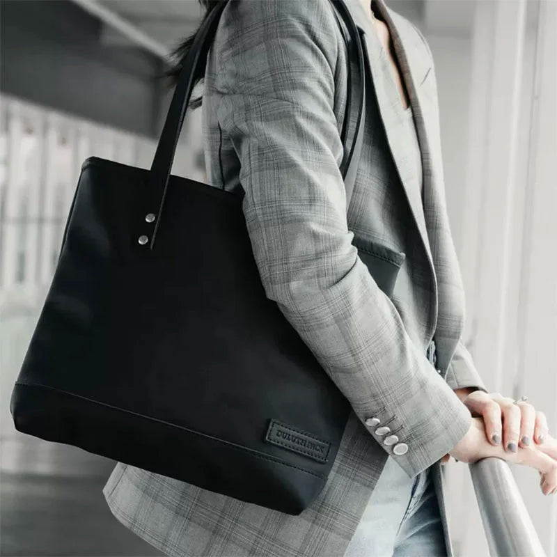 Superior Series Leather Lakewalk Tote in black premium leather with riveted handles, carried by a person in a gray plaid blazer near an industrial window setting.