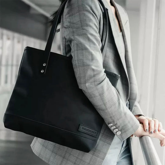Superior Series Leather Lakewalk Tote in black premium leather with riveted handles, carried by a person in a gray plaid blazer near an industrial window setting.