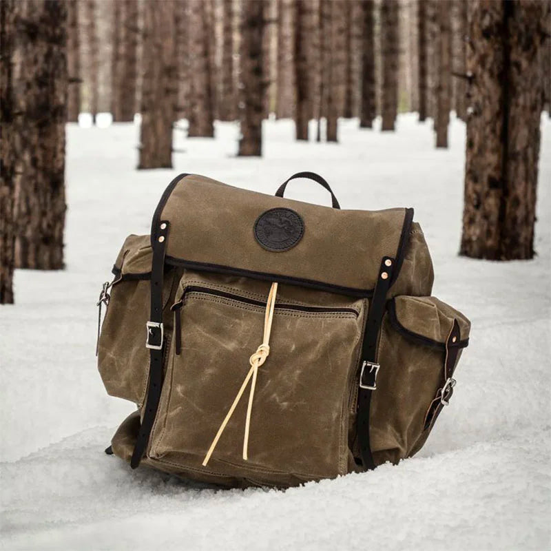 Rambler backpack in olive drab waxed canvas with black leather straps, designed for rugged outdoor use, sitting on snow in a pine forest setting.