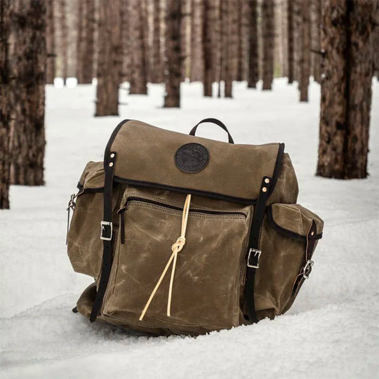 Rambler backpack in olive drab waxed canvas with black leather straps, designed for rugged outdoor use, sitting on snow in a pine forest setting.