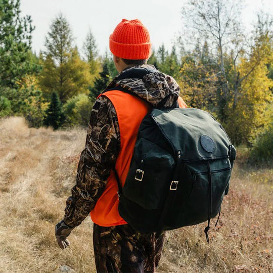 Rambler black canvas backpack worn by a hiker in bright orange and camo clothing walking through a forest trail in autumn wilderness.