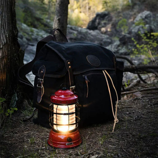 Rambler black canvas backpack with leather accents beside a glowing lantern in a rugged outdoor forest setting, showcasing durable handmade craftsmanship.