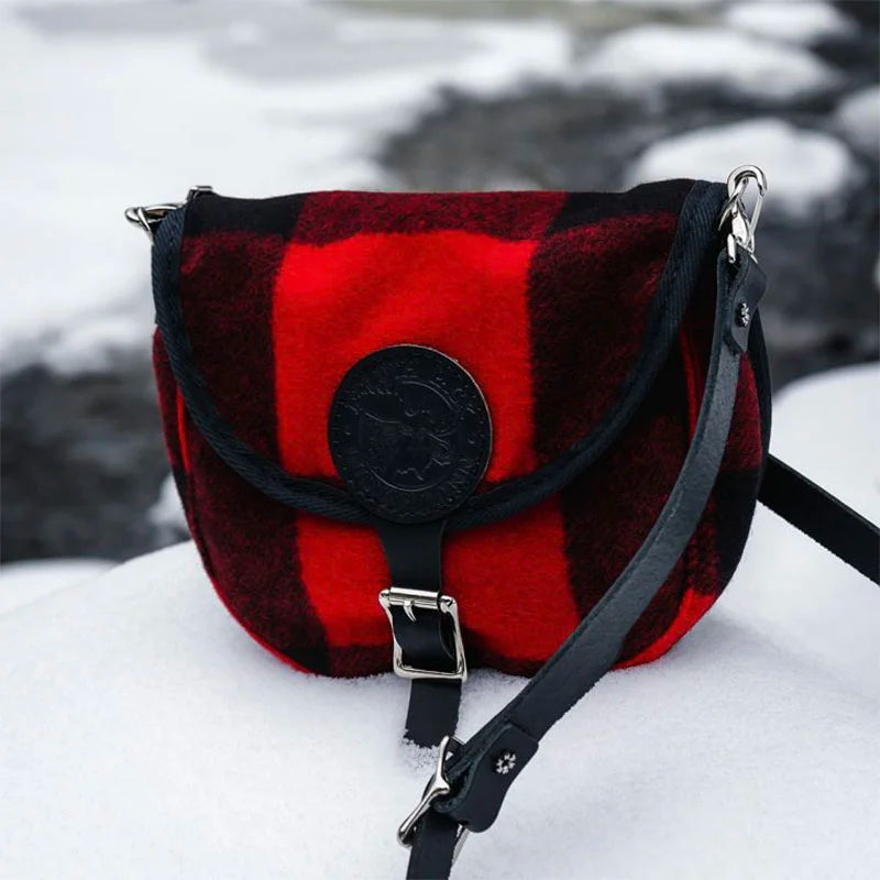 Classic Wool Small Shell Purse in red and black wool plaid with leather strap, resting on snow near a cold outdoor stream setting