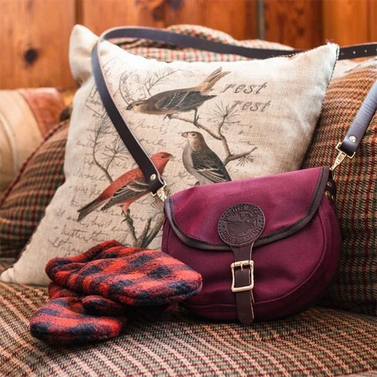 Small Shell Purse in burgundy with adjustable leather strap resting on a patterned fabric chair next to plaid gloves and a bird-illustrated pillow in a cozy indoor setting.
