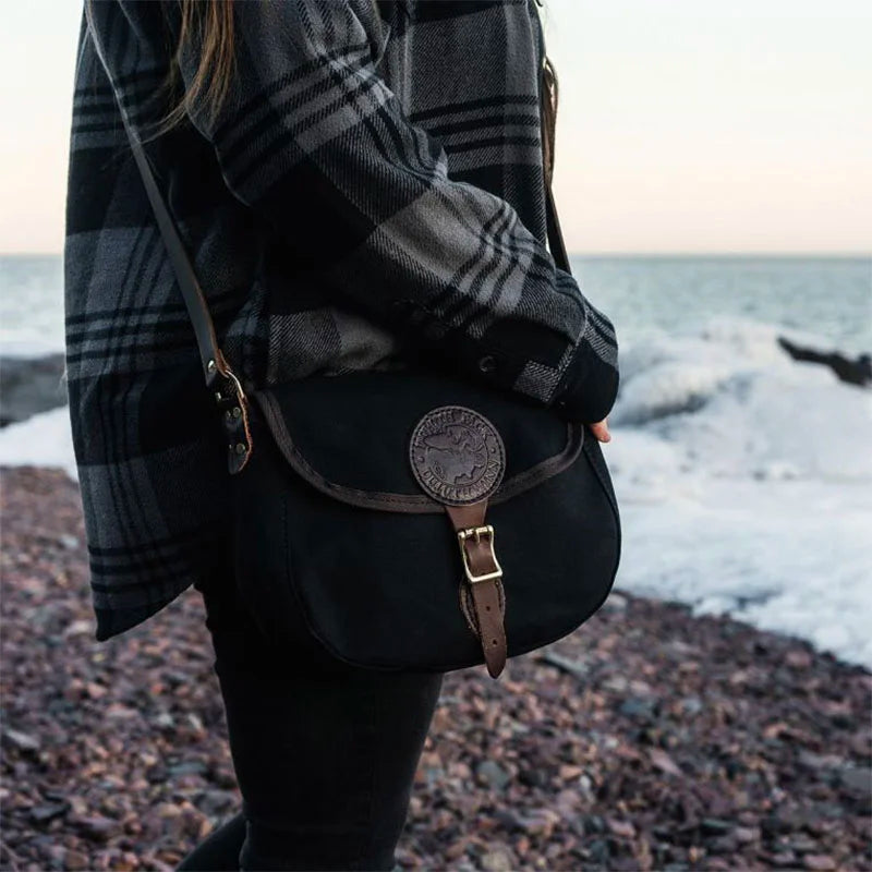 Medium Shell Purse in black with leather accents by Duluth Pack, worn outdoors on a rocky shoreline showcasing rugged, handmade craftsmanship and heritage quality.