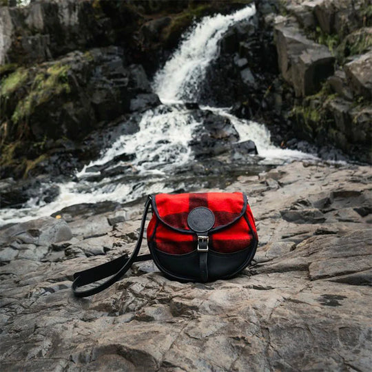 Deluxe Medium Classic Wool Shell Purse with timeless red wool exterior and leather strap, showcased on rugged rocks near a flowing waterfall in an outdoor setting.