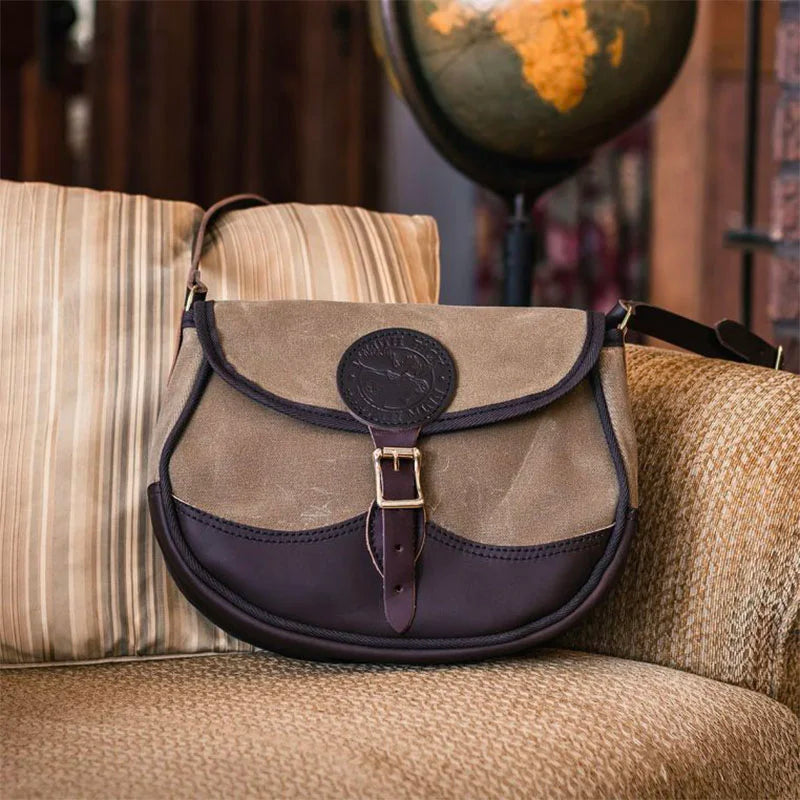 Deluxe Medium Shell Purse in waxed khaki canvas and dark leather, resting on a cozy armchair with a vintage globe in the background, showcasing rugged heritage style.