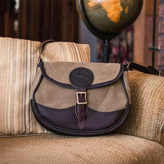 Deluxe Medium Shell Purse in waxed khaki canvas and dark leather, resting on a cozy armchair with a vintage globe in the background, showcasing rugged heritage style.