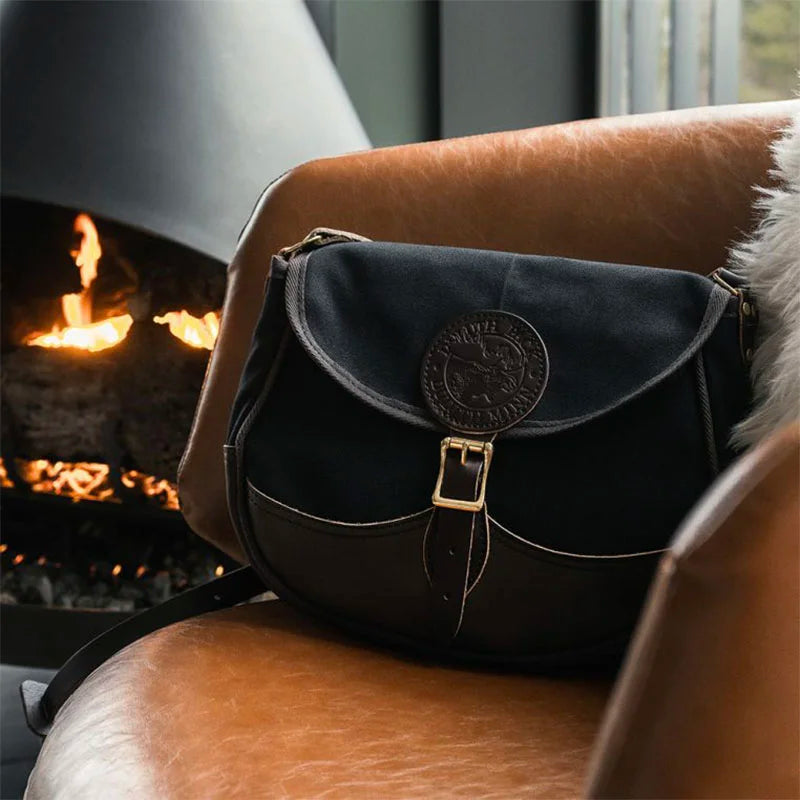 Deluxe Medium Shell Purse in black with leather accents by Duluth Pack, resting on a leather chair near a cozy fireplace in a rustic indoor setting.