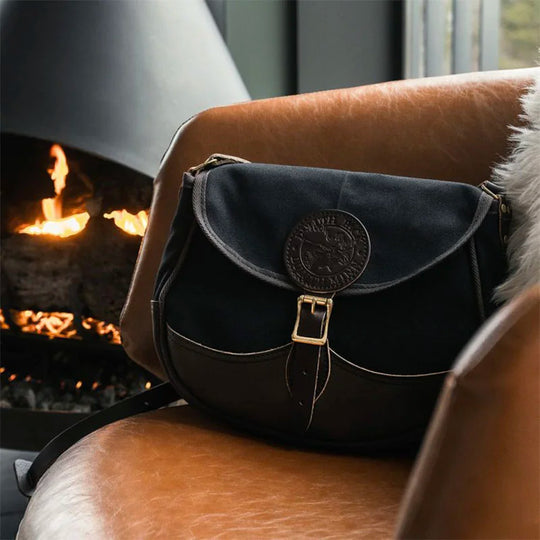 Deluxe Medium Shell Purse in black with leather accents by Duluth Pack, resting on a leather chair near a cozy fireplace in a rustic indoor setting.