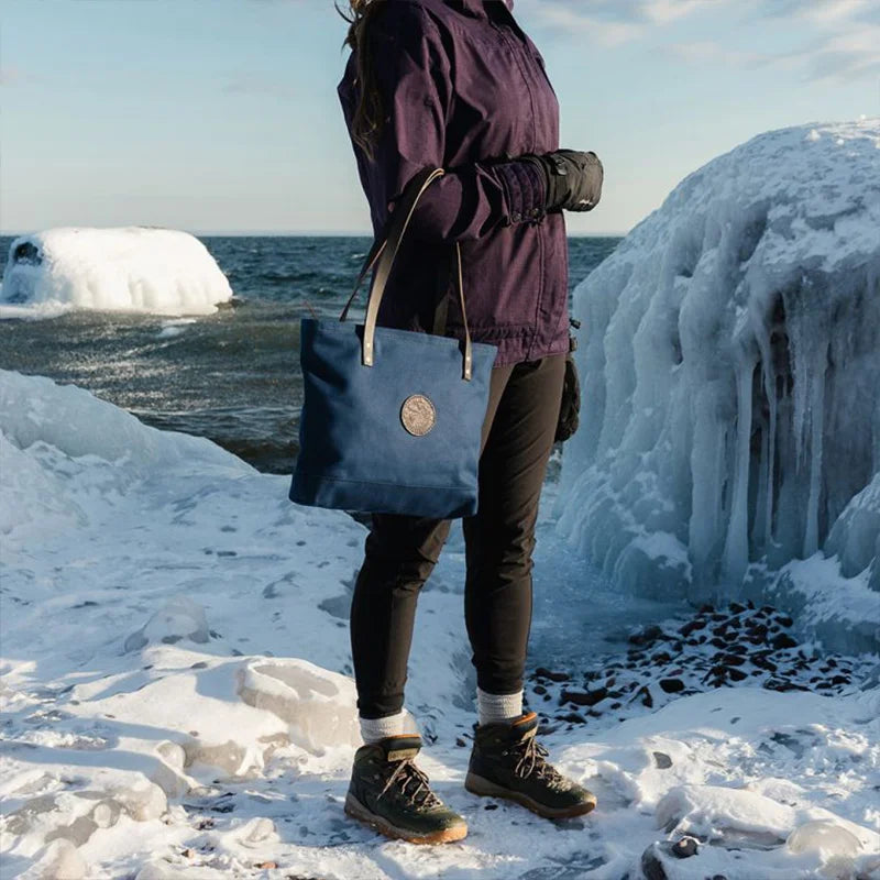 Lakewalk Tote in navy with riveted leather handles, carried outdoors on a snowy lakeshore, showcasing rugged, handmade canvas durability by Duluth Pack.