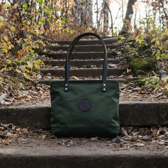 Lakewalk Tote in Olive Drab with riveted black leather handles, showcased outdoors on stone steps surrounded by autumn foliage, highlighting rugged craftsmanship.