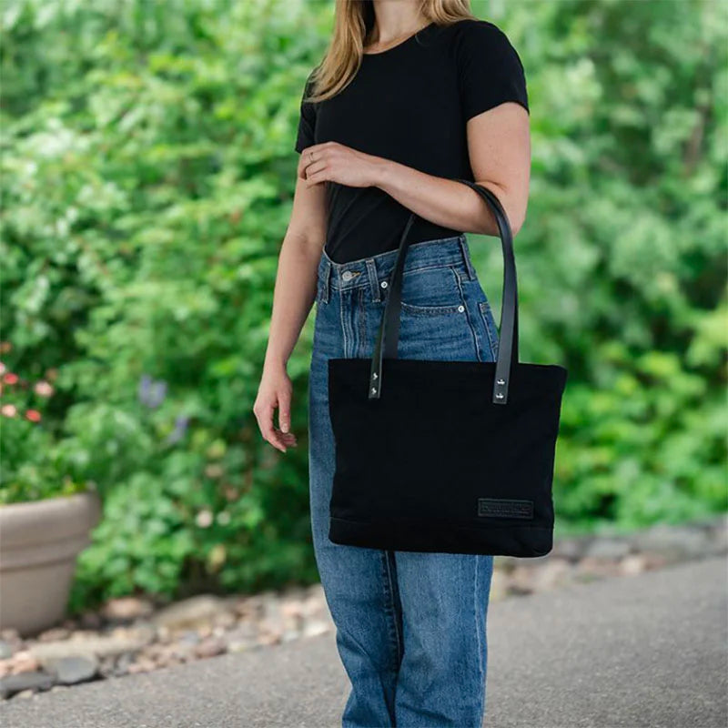 Superior Series Canvas Lakewalk Tote in black canvas with leather straps carried outdoors by a woman wearing casual jeans and a black shirt, highlighting its rugged durability.