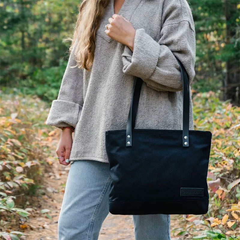 Superior Series Canvas Lakewalk Tote in black canvas with leather straps by Duluth Pack, carried outdoors on a forest trail during autumn.