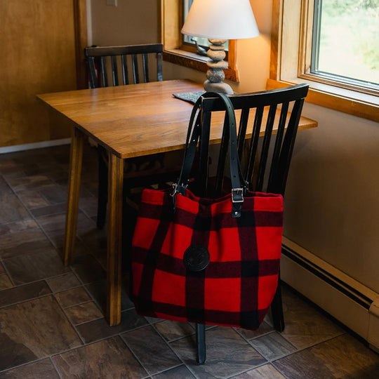 Classic Wool Market Tote in red and black plaid wool with adjustable leather handles hanging on a wooden chair in a warm, rustic indoor setting.