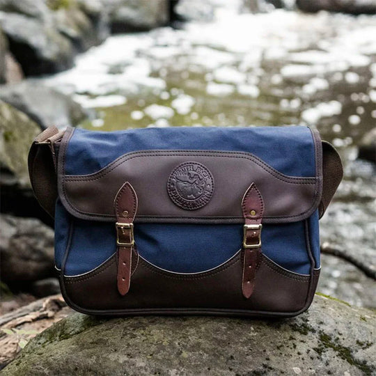 Deluxe Book Bag in navy with handcrafted leather trim and antique brass hardware, sitting on a rock by a flowing creek, showcasing rugged outdoor style.