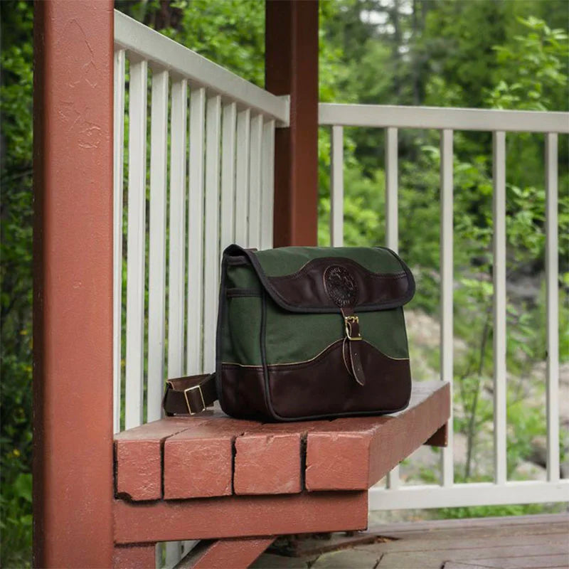 Field Satchel in rugged green 15-ounce canvas with thick premium leather detailing and comfortable web shoulder strap, displayed outdoors on rustic wooden railing.