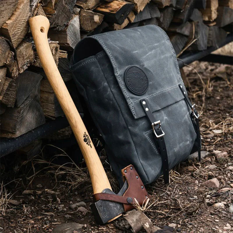 Ranger Pack in black rugged canvas with leather straps resting outdoors against stacked firewood beside a wooden-handled axe, showcasing durable outdoor gear.