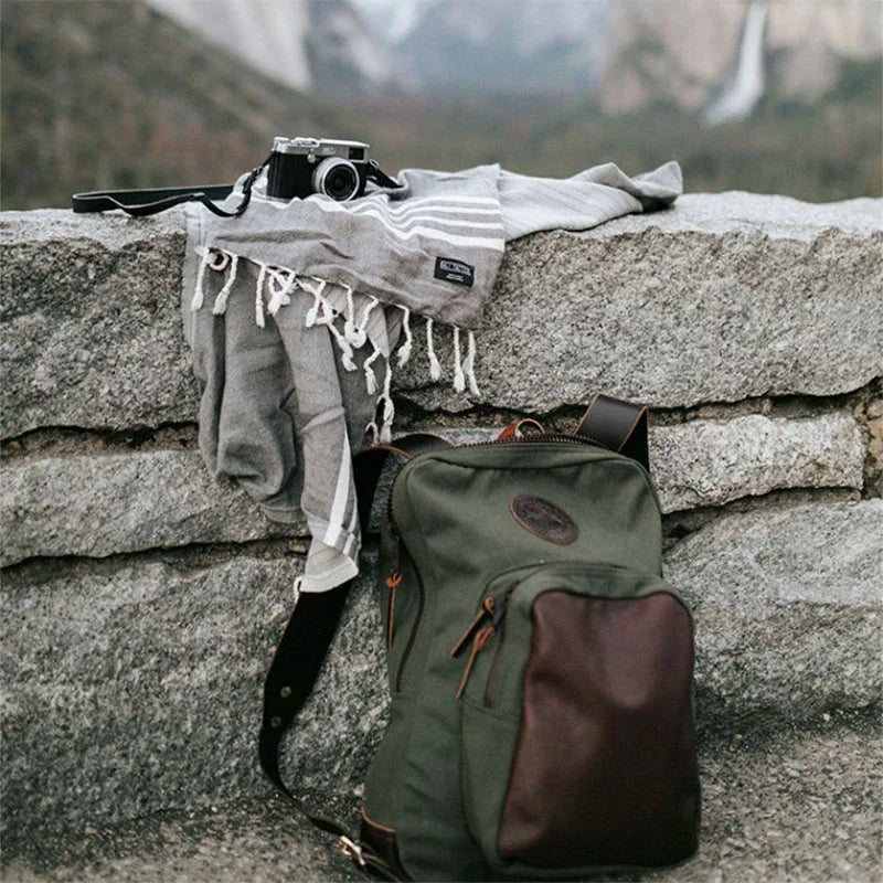Deluxe Laptop Backpack in Olive Drab featuring handmade canvas and leather, set outdoors on a stone wall with a vintage camera and wool scarf, by Duluth Pack.