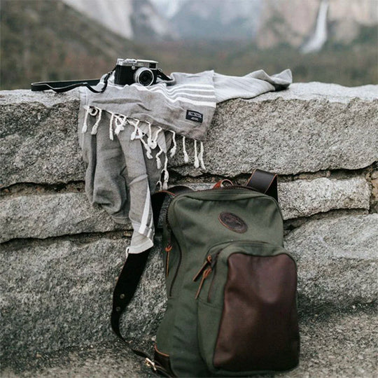 Deluxe Laptop Backpack in Olive Drab featuring handmade canvas and leather, set outdoors on a stone wall with a vintage camera and wool scarf, by Duluth Pack.