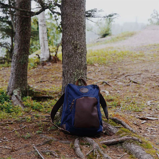 Deluxe Laptop Backpack in navy with brown leather pocket, made of rugged canvas, resting against a tree in a forest setting by Duluth Pack.
