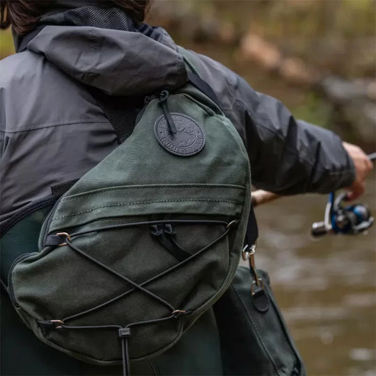 Sling Pack in Olive Drab crafted from durable handmade canvas, worn by an angler casting a fishing rod in an outdoor river setting.