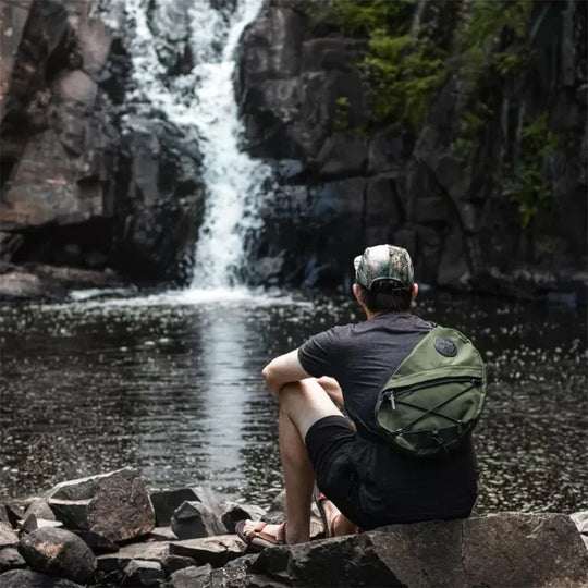 DOODLE BAG CHARM (PDP Test) in olive green canvas resting on a rugged outdoor backpack, worn by a man seated beside a rocky waterfall scene, reflecting durable heritage gear by DOUGLAS COMPANY, INC.