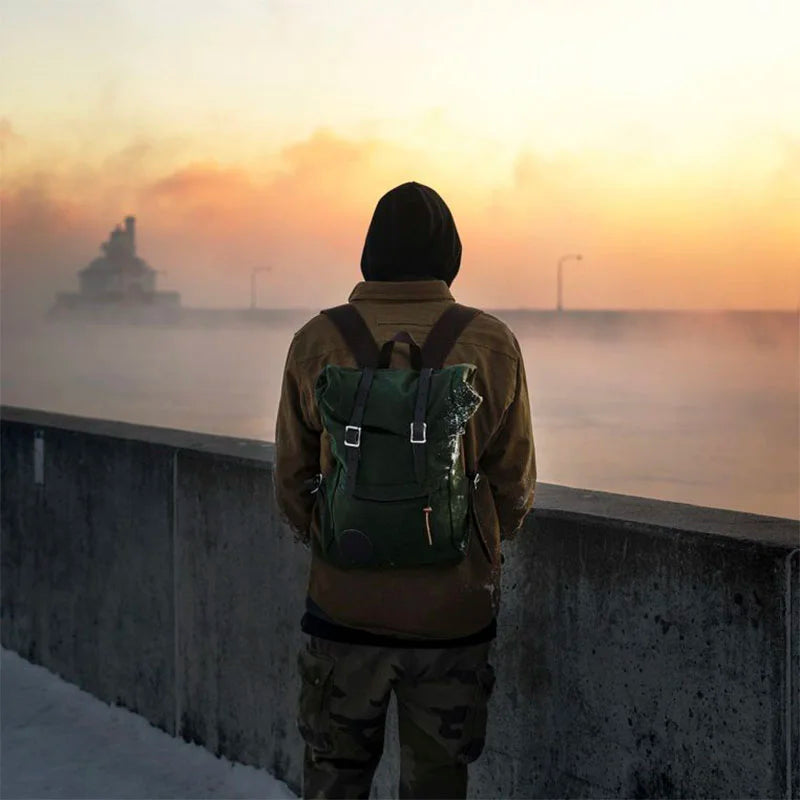 Roll-Top Scout Pack in dark green canvas with leather accents, worn by a person overlooking a foggy waterfront at sunrise, showcasing durable outdoor craftsmanship.