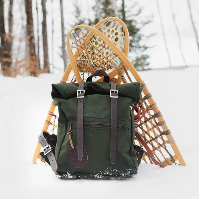 Roll-Top Scout Pack in dark green canvas with leather straps, positioned outdoors in snow against traditional wooden snowshoes, showcasing rugged outdoor gear craftsmanship.