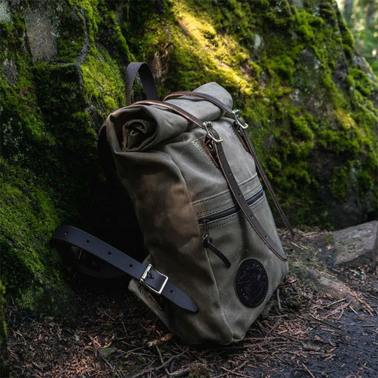 Roll-Top Scout Pack in olive canvas with leather straps by Duluth Pack, designed for durable outdoor use, shown resting against mossy rocks in a forest setting.