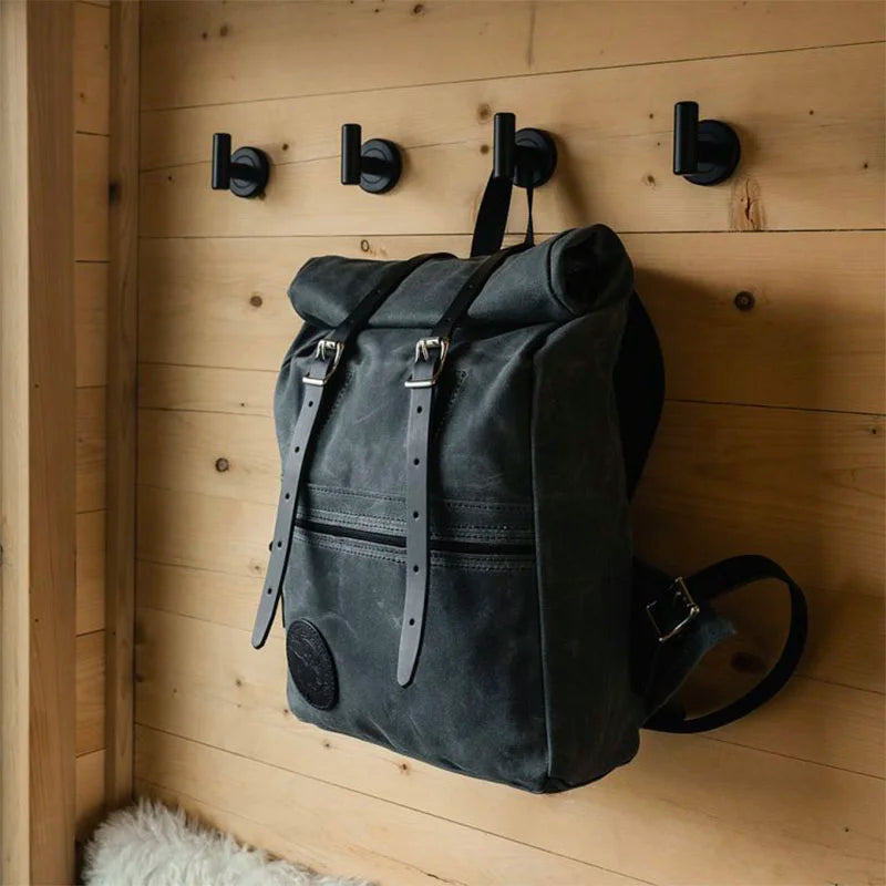 Roll-Top Scout Pack in dark canvas with leather straps hanging on wooden wall hooks, showcasing durable handmade outdoor gear craftsmanship.
