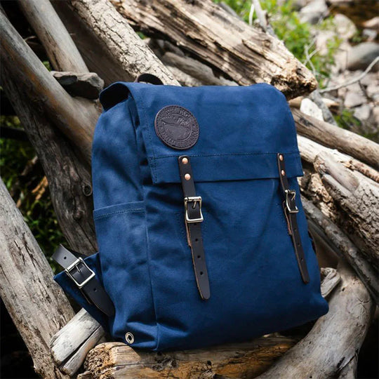 Laptop Scoutmaster in navy featuring handmade canvas and leather straps by Duluth Pack, set against a natural outdoor background with logs.