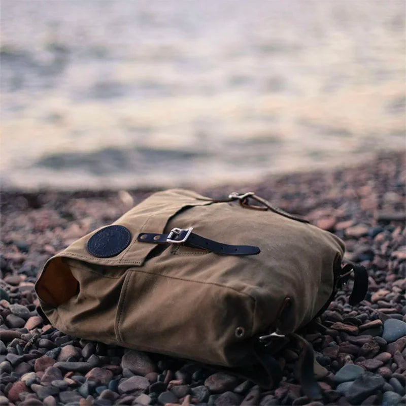 Laptop Scoutmaster in Waxed Khaki canvas with leather straps by Duluth Pack, shown resting on rocky terrain near a calm body of water, highlighting its rugged outdoor design and heritage craftsmanship.