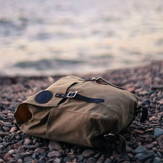 Laptop Scoutmaster in Waxed Khaki canvas with leather straps by Duluth Pack, shown resting on rocky terrain near a calm body of water, highlighting its rugged outdoor design and heritage craftsmanship.