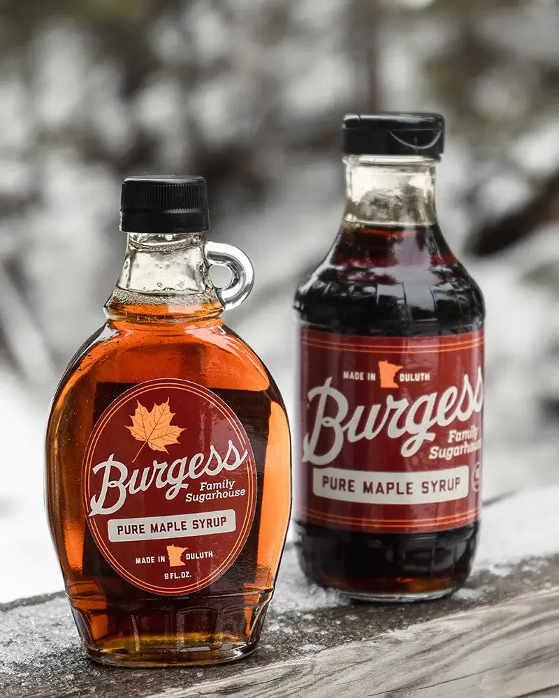 Two bottles of Burgess Family Sugarhouse maple syrup (one 8 Oz size and one 16 oz size) on a wooden surface with a blurred, snowy natural background.
