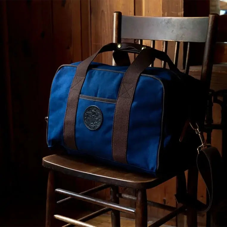 Small Safari Duffel In Navy Blue Sitting On A Rustic Wooden Chair In A Vintage Cabin