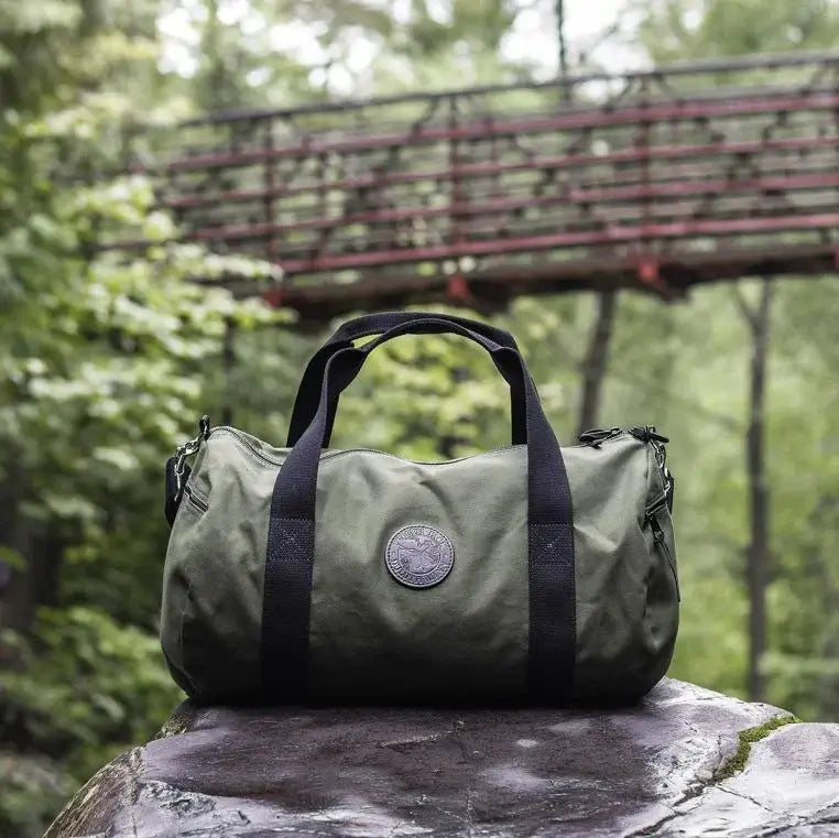 Round Duffel In Olive Drab Green Rests Under A Bridge In A Green Forrest