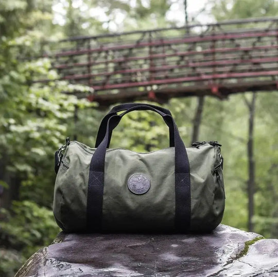 Round Duffel In Olive Drab Green Rests Under A Bridge In A Green Forrest