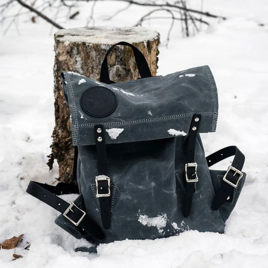 Scout Pack in waxed grey canvas with leather straps, resting on snow-covered ground against a tree stump, showcasing durable handmade outdoor craftsmanship.