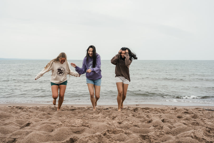 Three women wearing casual sweatshirts and shorts walking barefoot along a sandy beach with the ocean and cloudy sky in the background.