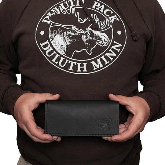 Leather Checkbook Cover in black premium leather with embossed logo, held by person wearing a Duluth Pack hoodie, showcasing durable handmade craftsmanship.
